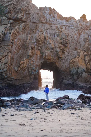 Person standing by a rock arch on a beach.
